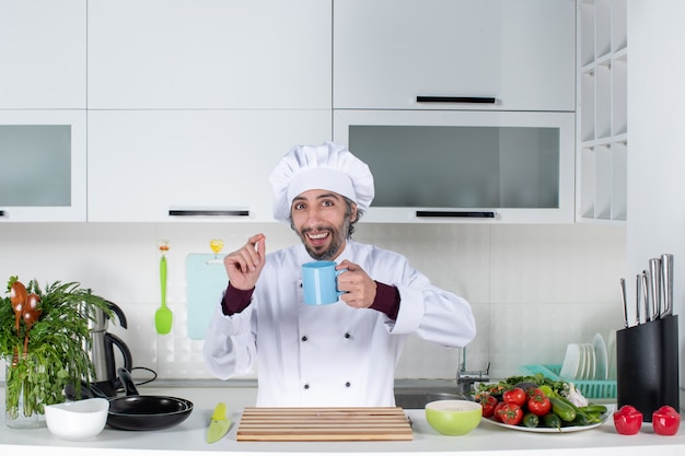 Vista frontal sonriente chef carismático en uniforme sosteniendo la taza de pie detrás de la mesa de la cocina