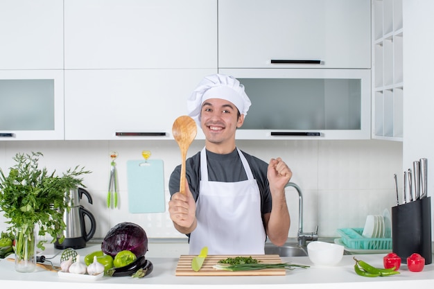 Vista frontal sonriente chef masculino en uniforme sosteniendo una cuchara de madera detrás de la mesa de la cocina