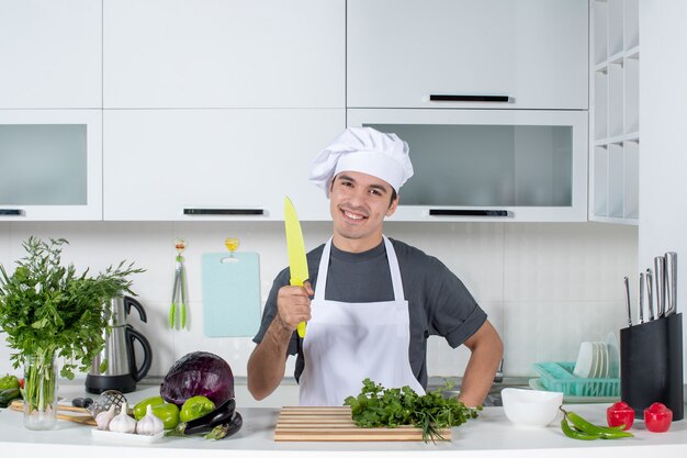 Vista frontal sonriente chef masculino en uniforme sosteniendo un cuchillo en la cocina