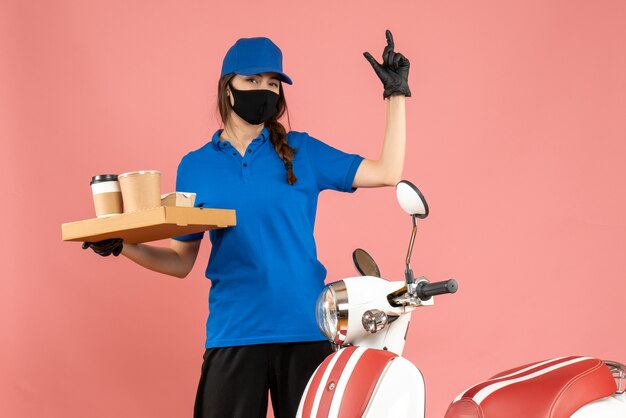 Vista frontal de la sonriente chica de mensajería con guantes de máscara médica de pie junto a la motocicleta sosteniendo pequeños pasteles de café sobre fondo de color melocotón pastel