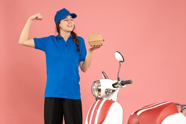 Vista frontal de la sonriente chica de mensajería de pie junto a la motocicleta sosteniendo la torta mostrando musculoso sobre fondo de color melocotón pastel