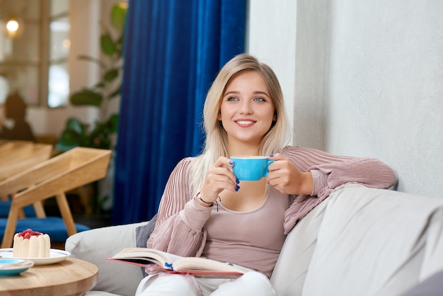 Vista frontal de la sonriente chica rubia bebiendo un sabroso café sentado en el sofá blanco