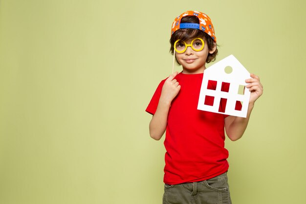 Una vista frontal sonriente chico lindo en camiseta roja con papel de la casa en el espacio de color piedra