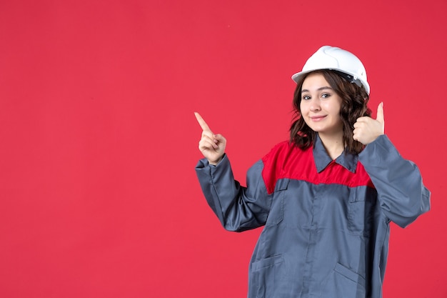 Vista frontal de la sonriente constructora confiada en uniforme con casco y haciendo gesto de ok apuntando hacia arriba sobre fondo rojo aislado