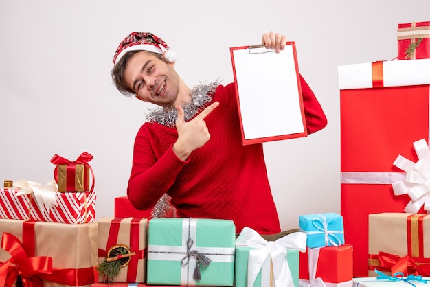 Vista frontal sonriente joven apuntando con el dedo portapapeles sentado alrededor de regalos de navidad