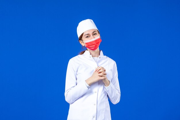 Vista frontal sonriente joven enfermera en traje médico con máscara protectora roja sobre azul