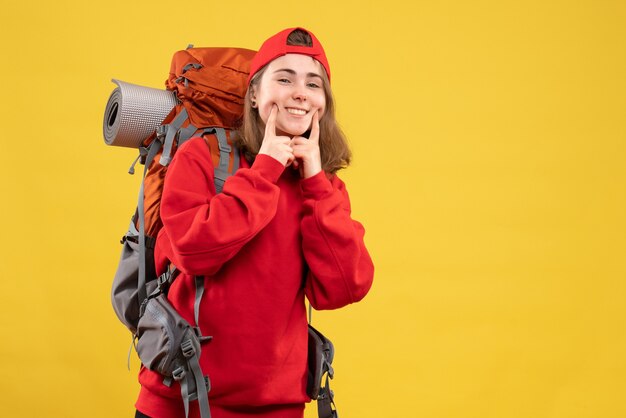 Vista frontal sonriente joven turista con mochila y gorra roja poniendo el dedo en la mejilla
