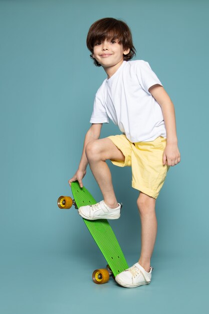 Una vista frontal sonriente lindo niño montando patineta verde en camiseta blanca y pantalones cortos naranjas en el espacio azul