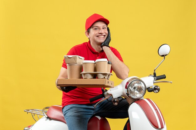 Vista frontal del sonriente mensajero esperanzado hombre vestido con blusa roja y guantes de sombrero en máscara médica entregando orden sentado en scooter con órdenes