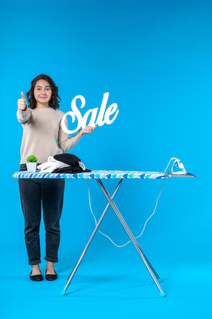 Vista frontal de la sonriente mujer joven feliz de pie detrás de la tabla de planchar y mostrando el icono de venta haciendo un gesto de ok en la pared azul