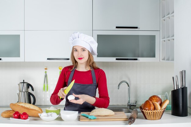 Vista frontal sonriente mujer rubia con sombrero de cocinero y delantal rompiendo un huevo con un cuchillo en la cocina