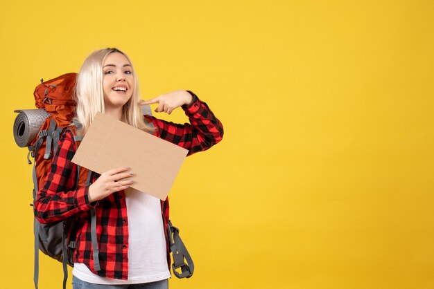 Vista frontal sonriente mujer rubia viajera con su mochila sosteniendo cartón de pie en la pared amarilla