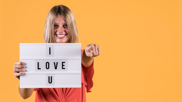 Vista frontal sonriente mujer sosteniendo caja de luz con te amo texto contra superficie amarilla