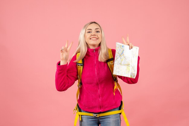 Vista frontal sonriente mujer viajera con mochila amarilla sosteniendo el mapa gesticulando el signo de la victoria