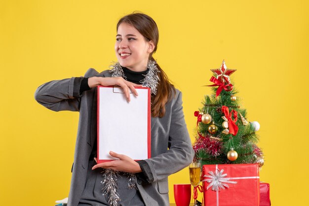Vista frontal sonriente niña sosteniendo documentos de pie cerca del árbol de Navidad y cóctel de regalos