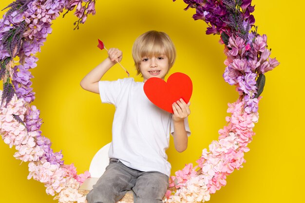 Una vista frontal sonriente niño rubio en camiseta blanca con forma de corazón sentado en la flor hecha soporte en el piso amarillo