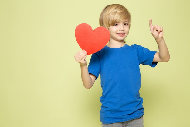 Una vista frontal sonriente niño rubio con forma de corazón en camiseta azul en el espacio de color piedra