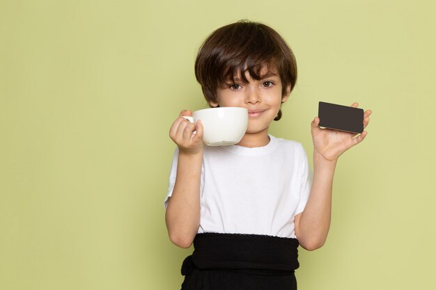 Una vista frontal sonriente niño con tarjeta negra y taza blanca en el escritorio de color piedra