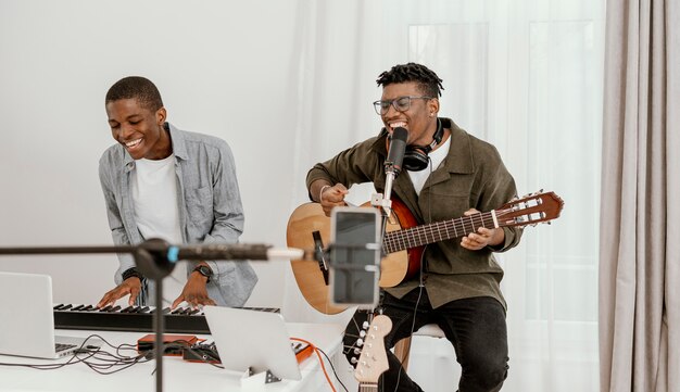 Vista frontal de sonrientes músicos masculinos en casa tocando la guitarra y el teclado eléctrico