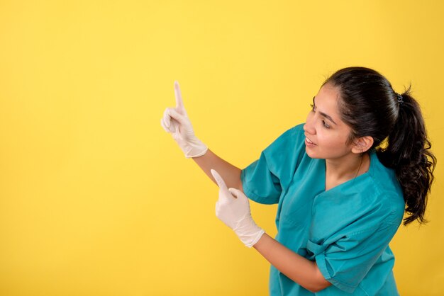 Vista frontal sonrió doctora con guantes de látex apuntando con el dedo hacia arriba sobre fondo amarillo