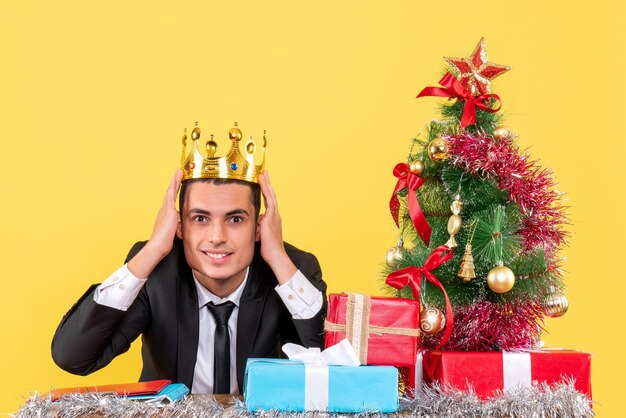 Vista frontal sonrió a hombre con corona sentado en la mesa árbol de Navidad y regalos