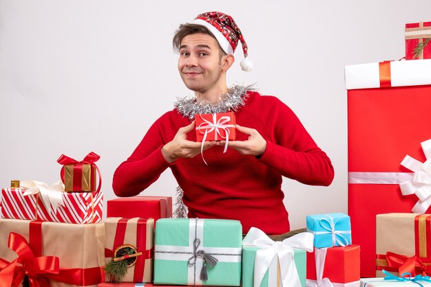 Vista frontal sonrió a joven con gorro de Papá Noel sentado alrededor de regalos de Navidad