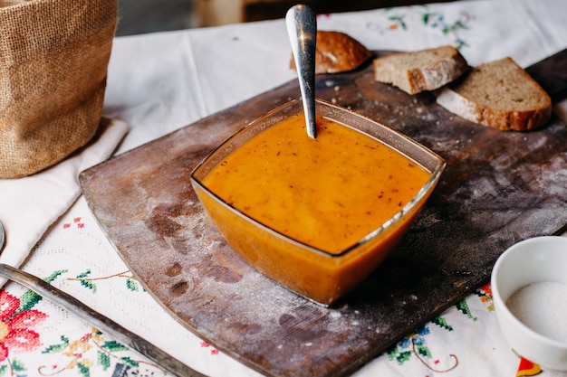 Una vista frontal de sopa de pimienta naranja dentro de un plato salado sabroso con una cuchara junto con panes de pan líquido de comida en el escritorio marrón