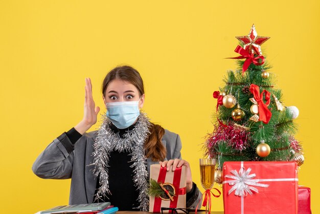 Vista frontal sorprendida chica con máscara médica sentada en la mesa levantando su mano árbol de Navidad y cóctel de regalos