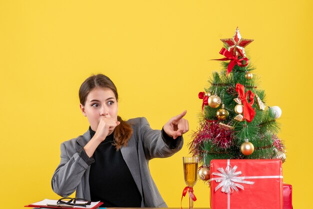 Vista frontal sorprendida niña sentada en el escritorio apuntando con el dedo algo árbol de Navidad y regalos cóctel