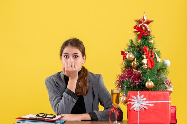 Vista frontal sorprendida niña sentada en el escritorio puso la mano a la boca árbol de Navidad y cóctel de regalos