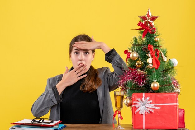 Vista frontal sorprendida niña sentada en la mesa cerca del árbol de Navidad y cóctel de regalos