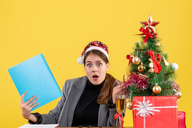 Vista frontal sorprendida niña con sombrero de Navidad sentado en la mesa árbol de Navidad y cóctel de regalos