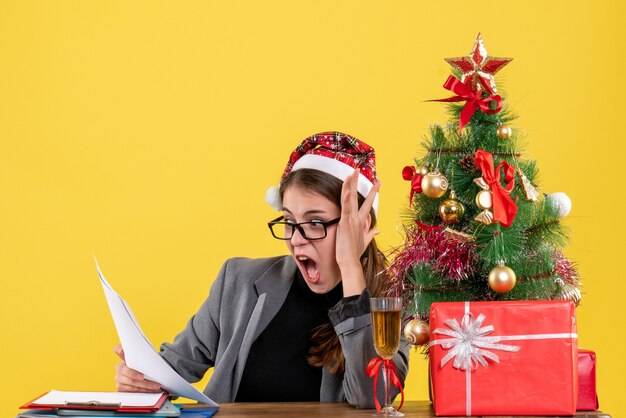 Vista frontal sorprendida niña con sombrero de Navidad sentado en la mesa mirando papeles árbol de Navidad y cóctel de regalos