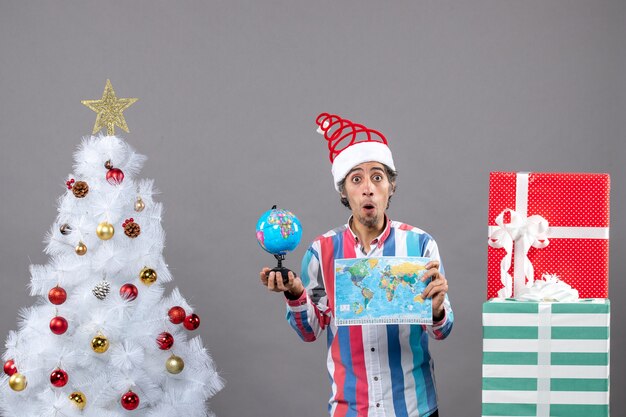 Vista frontal sorprendido hombre sosteniendo mapa del mundo y globo de pie cerca del árbol de Navidad blanco con estrella