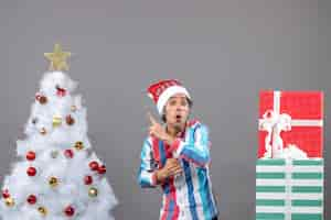 Foto gratuita vista frontal sorprendido joven con gorro de papá noel de resorte espiral mostrando árbol de navidad blanco