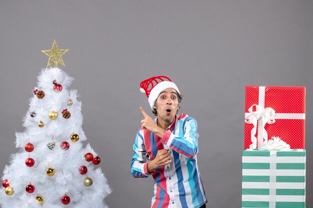 Vista frontal sorprendido joven con gorro de Papá Noel de resorte espiral mostrando árbol de Navidad blanco