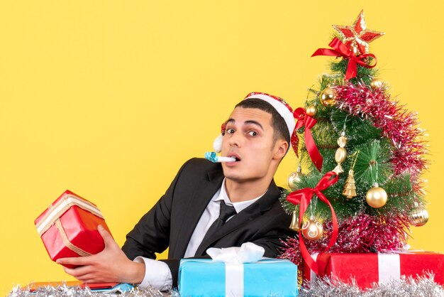 Vista frontal sorprendió al hombre con gorro de Papá Noel sentado en la mesa sosteniendo el presente usando el árbol de Navidad y los regalos de matraca