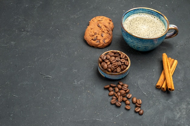 Vista frontal de una taza de café con semillas de café, palitos de canela, galletas en lugar oscuro y libre