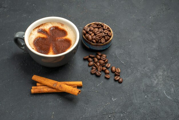 Vista frontal de una taza de café con semillas de café, palitos de canela en un lugar oscuro y libre