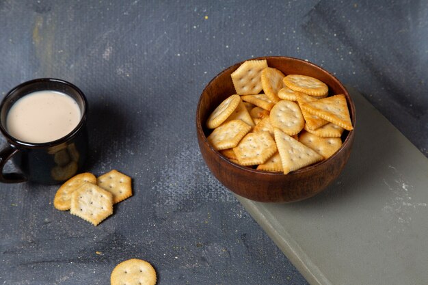 Vista frontal de la taza de leche con plato marrón lleno de galletas en la superficie gris