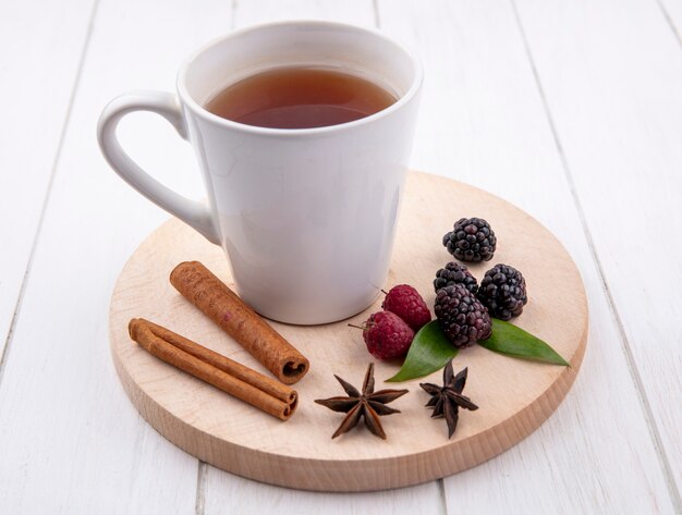 Vista frontal de la taza de té con canela, frambuesas y moras sobre un soporte sobre una superficie blanca