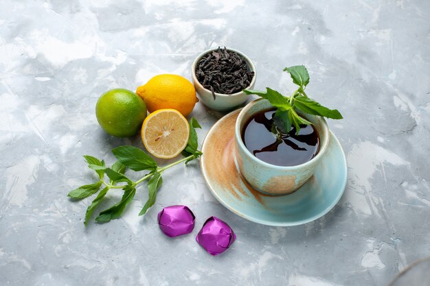 Vista frontal taza de té con limones frescos caramelos y té seco en la mesa de luz, color cítrico de frutas de té