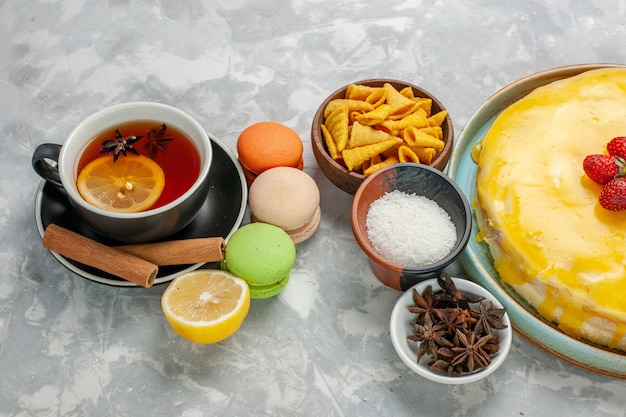 Vista frontal taza de té con macarons franceses y delicioso pastel de sirope amarillo sobre superficie blanca