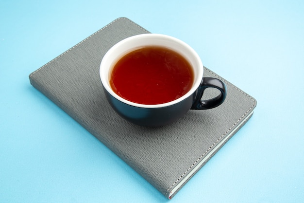 Vista frontal de una taza de té negro en el cuaderno gris sobre superficie azul