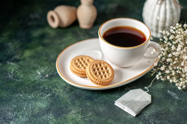Vista frontal de una taza de té con pequeñas galletas dulces en un plato blanco sobre la superficie oscura ceremonia de color pan de desayuno de vidrio beber azúcar