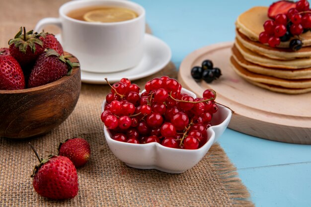 Vista frontal de la taza de té con una rodaja de limón y panqueques con grosellas rojas y negras y fresas sobre una superficie azul