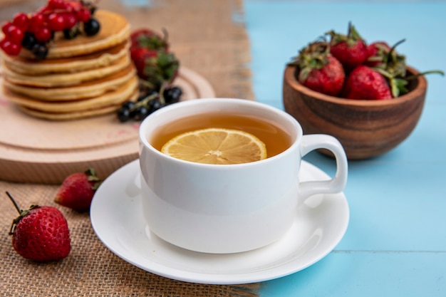 Vista frontal de la taza de té con una rodaja de limón y panqueques con grosellas rojas y negras y fresas sobre una superficie azul