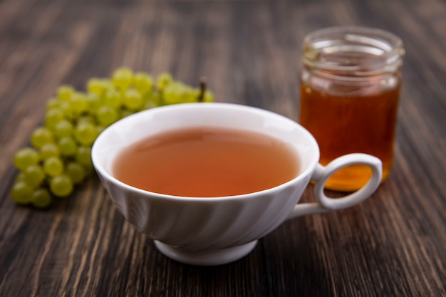 Vista frontal de una taza de té con uvas verdes y miel en un frasco sobre fondo de madera