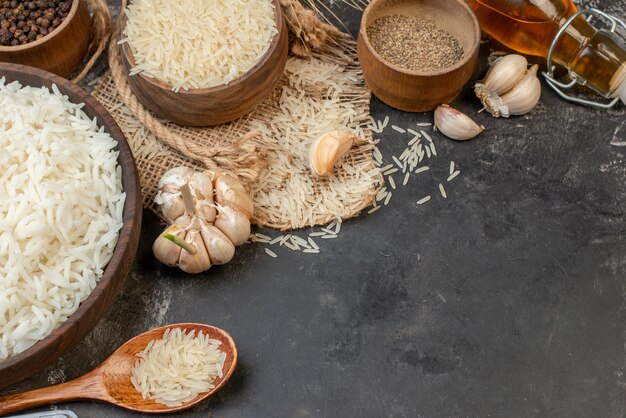 Vista frontal de tazones largos perfectos de arroz marrón sobre una toalla de color desnudo, una botella de aceite caída, ajos, pimientos sobre una mesa oscura