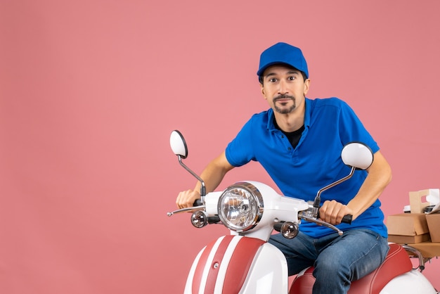 Vista frontal del tipo de mensajero feliz con sombrero sentado en scooter sobre fondo de melocotón pastel